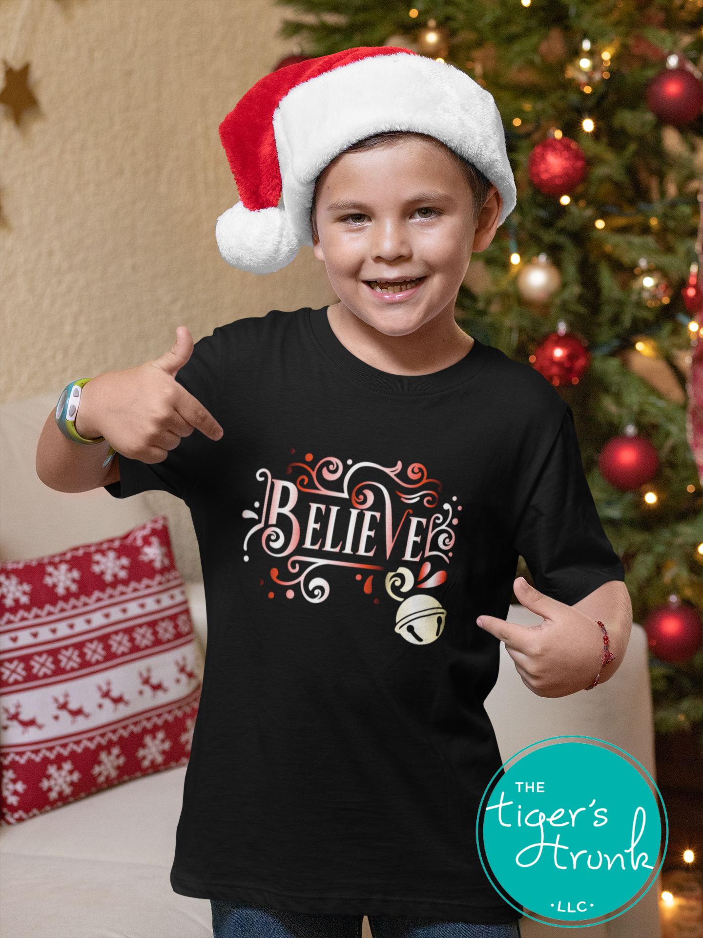 Child wearing a black t-shirt with 'BELIEVE' and a Christmas light design, pointing towards the camera, with a decorated Christmas tree in the background.