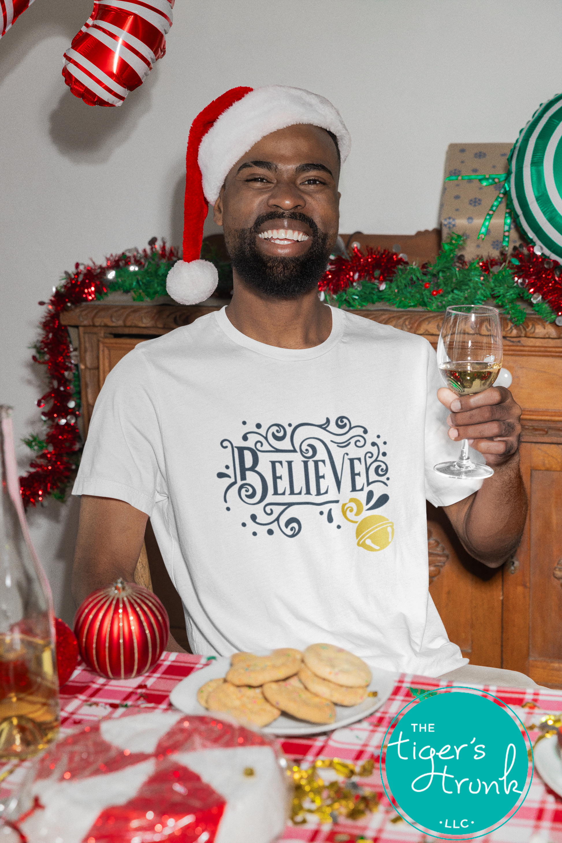 Man wearing a Santa hat and 'Believe' shirt holding a glass of wine at a festive table.
