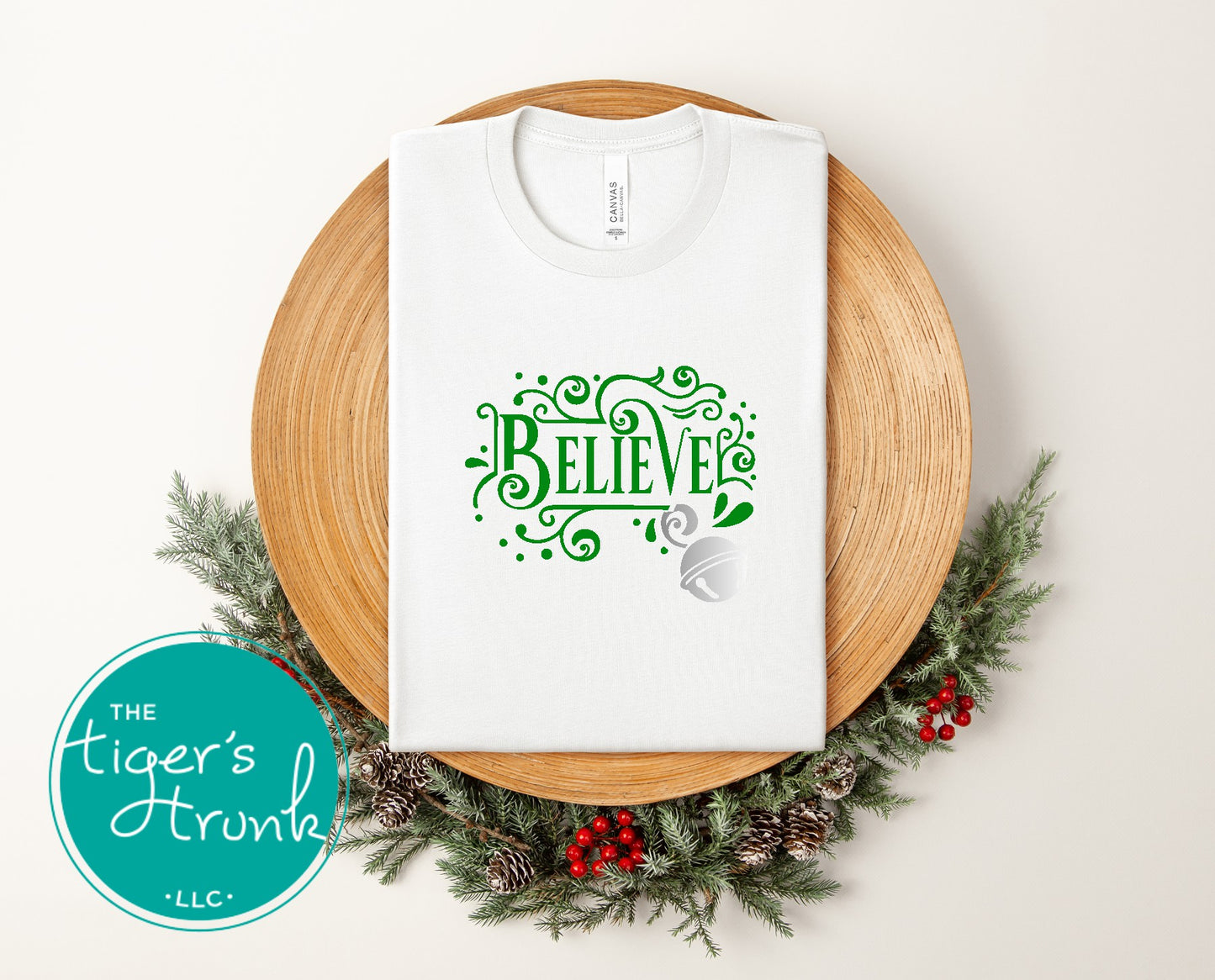White t-shirt with 'BELIEVE' design on a wooden plate surrounded by Christmas decorations, featuring The Tiger's Den logo.