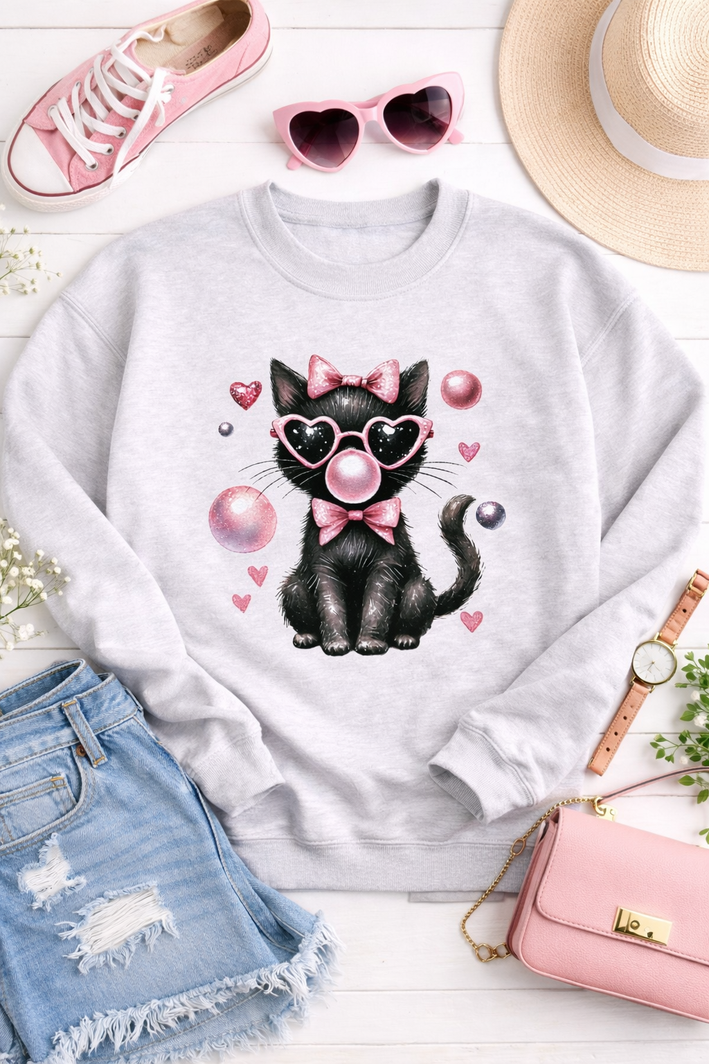 White sweatshirt with a black cat design wearing sunglasses and a bow, surrounded by accessories on a white surface.