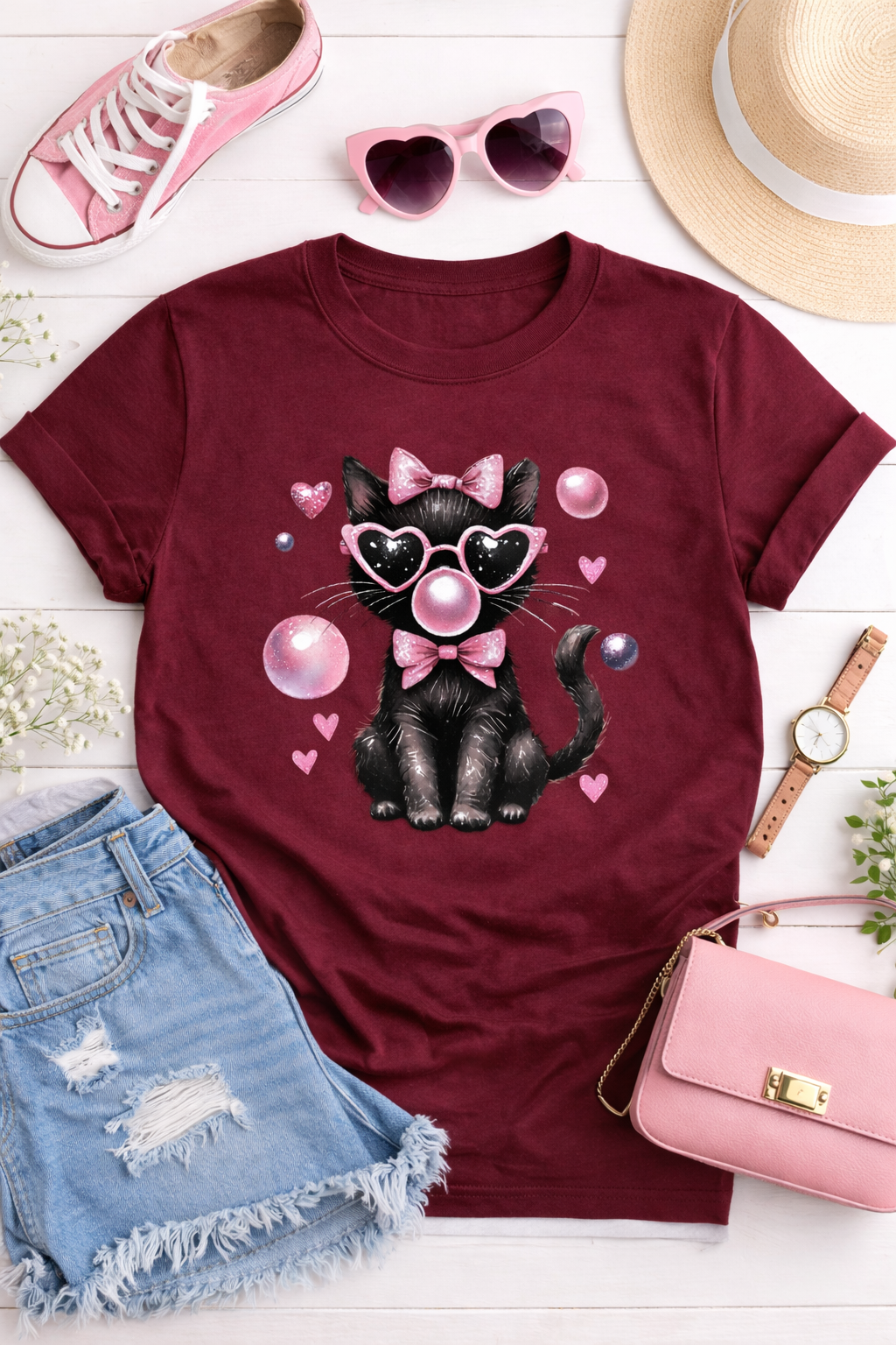 Maroon t-shirt with a black cat graphic, sunglasses, and pink accessories on a white surface.