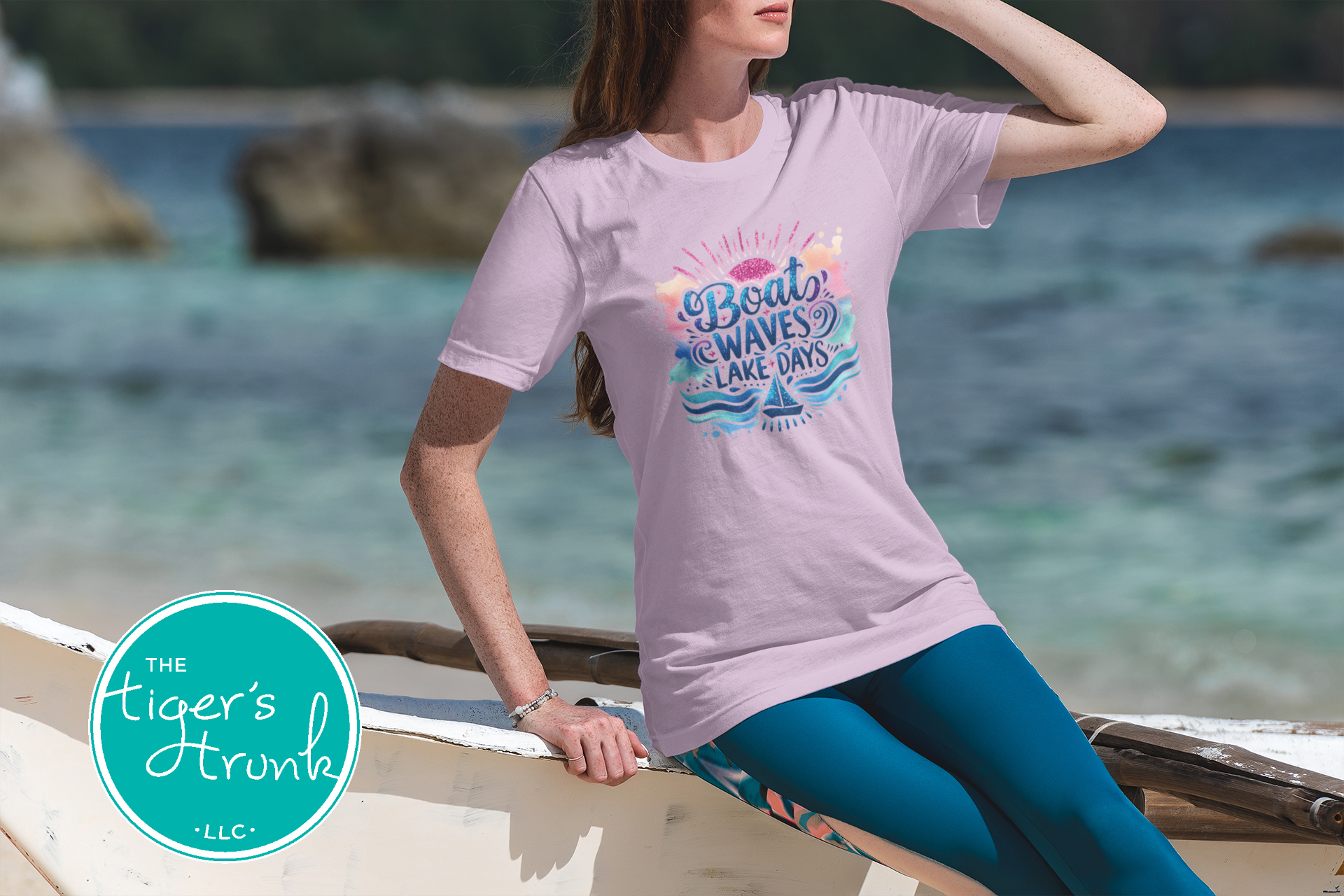 Woman wearing a light purple t-shirt with a graphic design sitting on a wooden bench by the water.