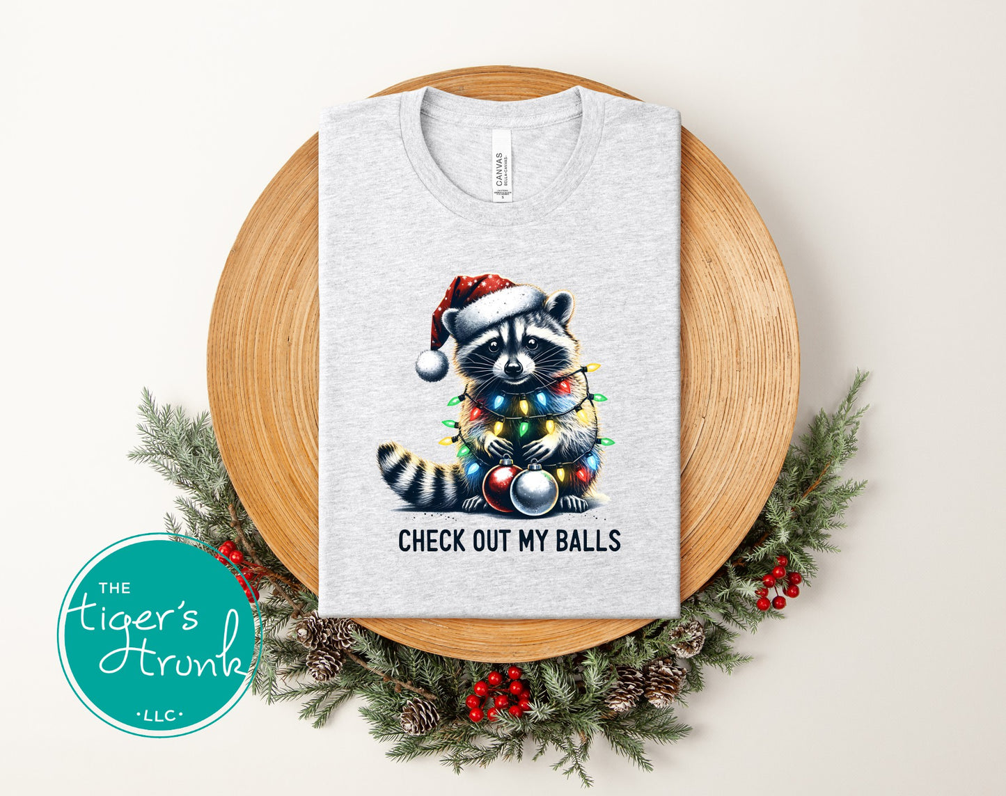 Gray t-shirt with a raccoon wearing a Santa hat and Christmas lights design, placed on a wooden board with a wreath.