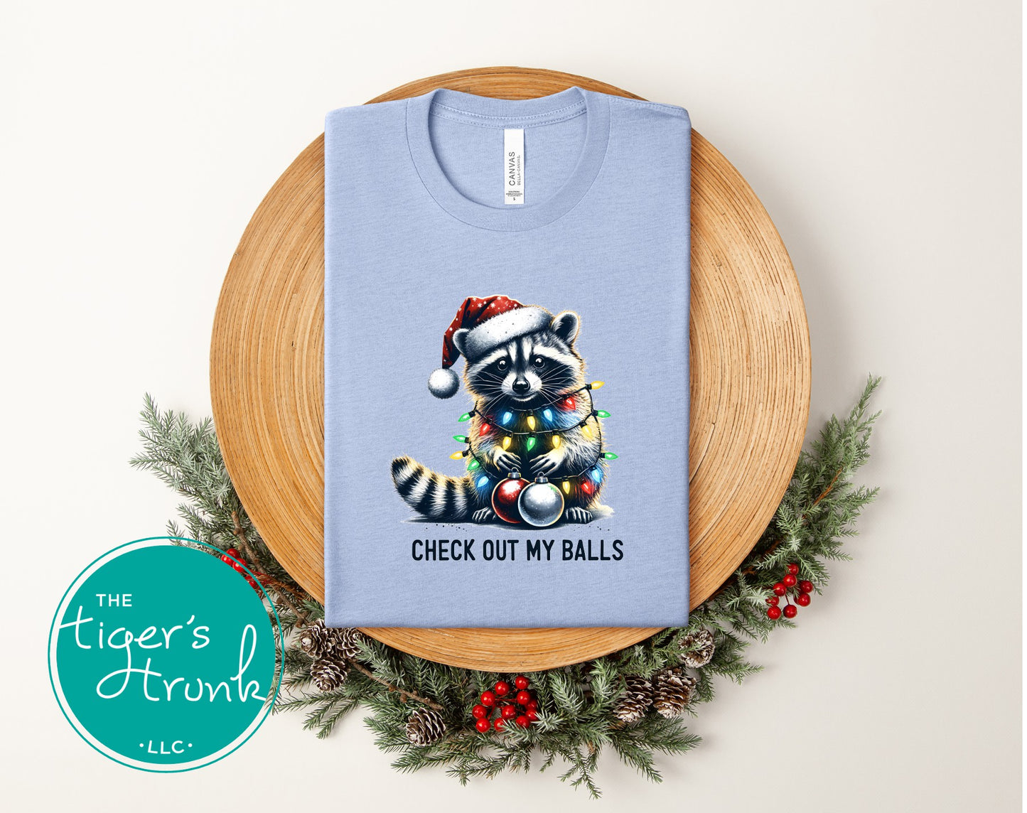 Light blue t-shirt with a Christmas-themed raccoon graphic on a wooden plate with greenery.