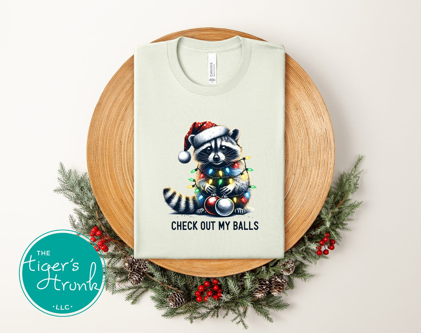 Light green t-shirt with a raccoon wearing a Santa hat and Christmas lights design, placed on a wooden plate with a wreath.