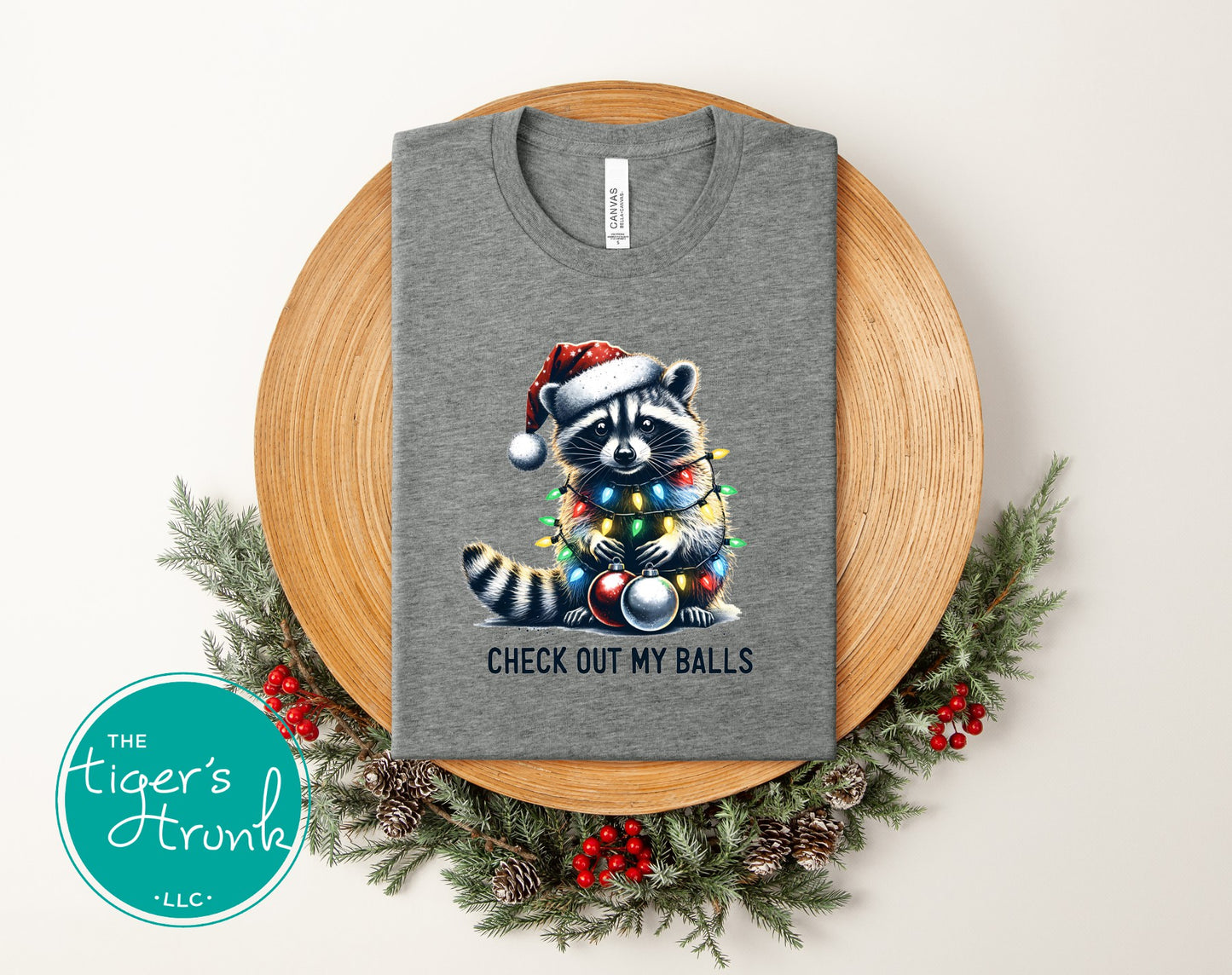 Gray t-shirt with a raccoon illustration and text on a wooden board with Christmas decorations.