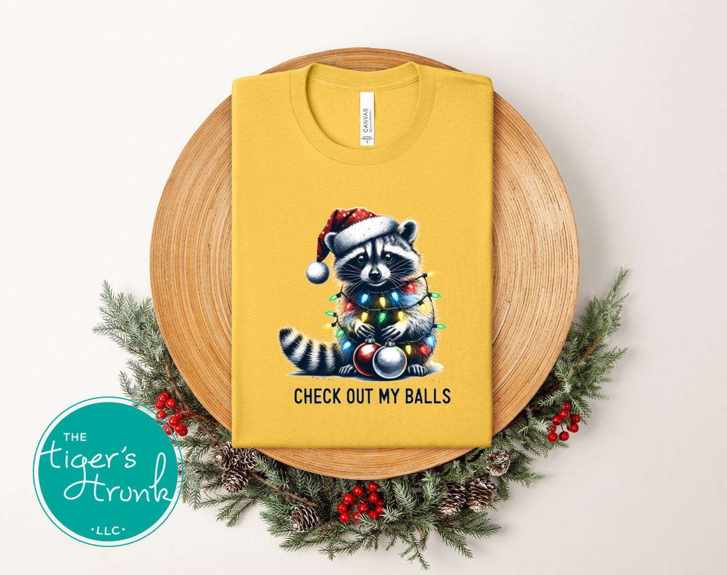 Yellow t-shirt with a raccoon wearing a Santa hat and Christmas lights design, placed on a wooden board with decorative elements.