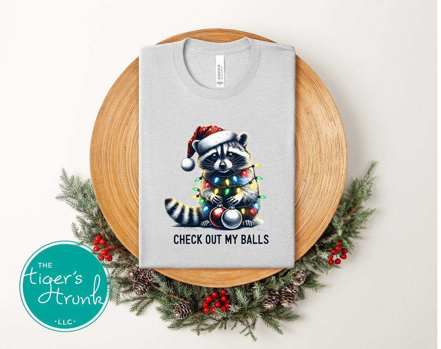 Gray t-shirt with a Christmas-themed raccoon design on a wooden plate with greenery, featuring 'The Tiger's Trunk' logo.