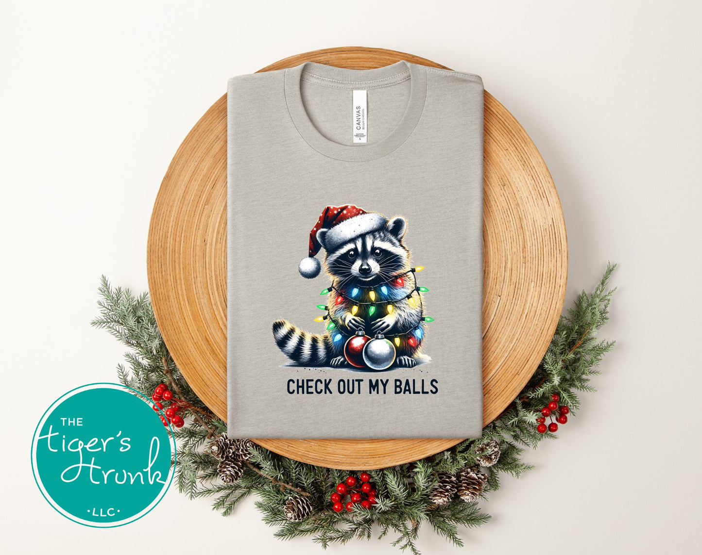 Gray t-shirt with a raccoon wearing a Santa hat and Christmas lights design, placed on a wooden board with decorative elements.