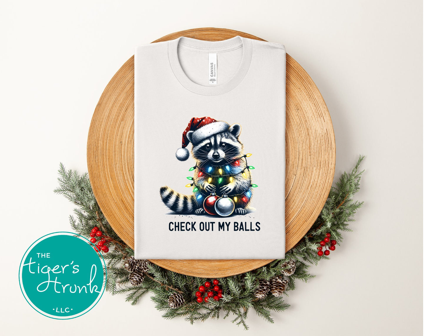 White t-shirt with a raccoon wearing a Santa hat and Christmas lights design, placed on a wooden board with decorative elements.