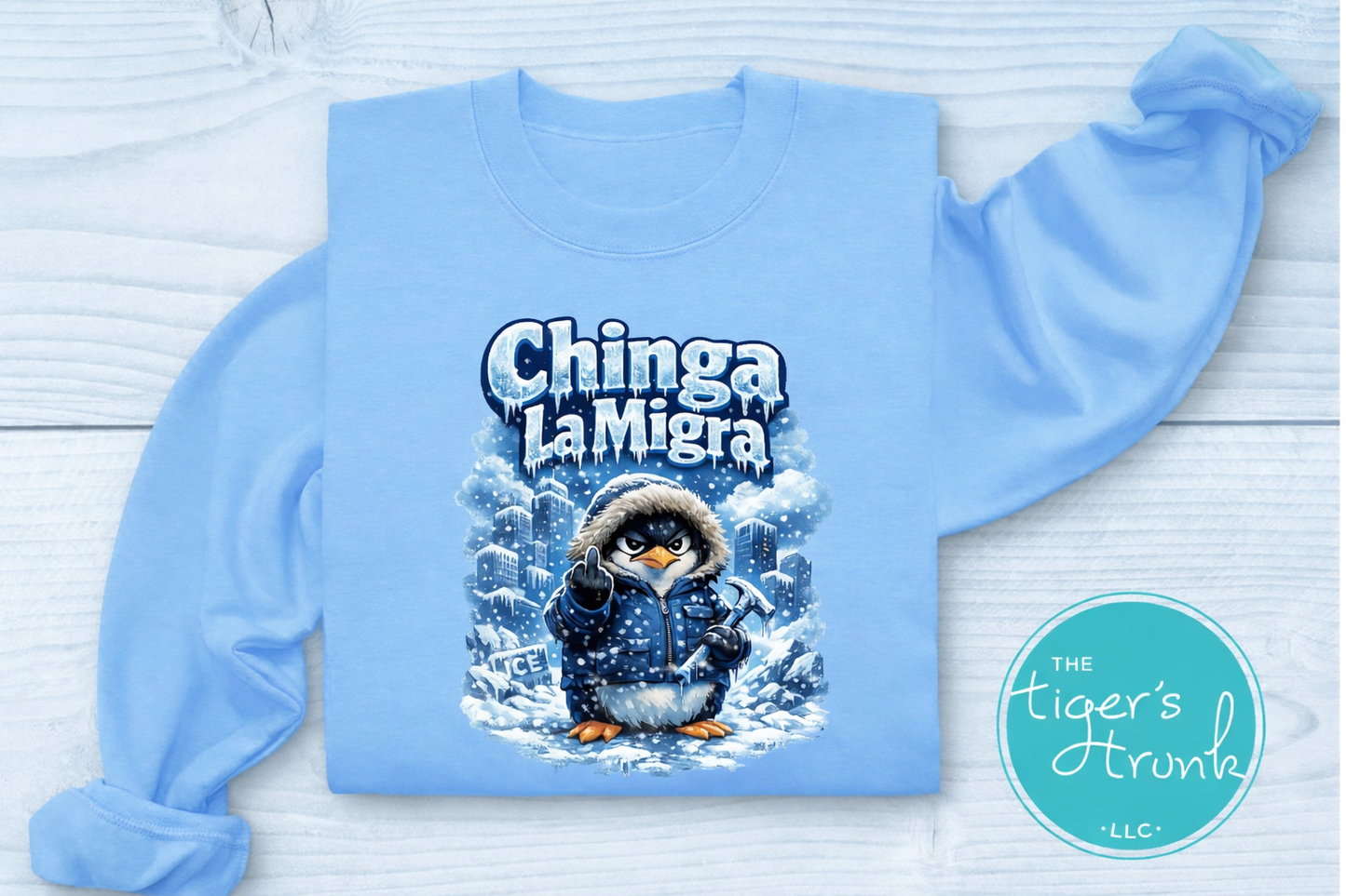 Light blue sweatshirt with a penguin graphic and 'Chinga La Migra' text on a white wooden background.