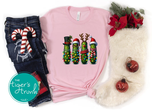 Pink t-shirt with Christmas pickle design, jeans, and holiday decorations on a white background.