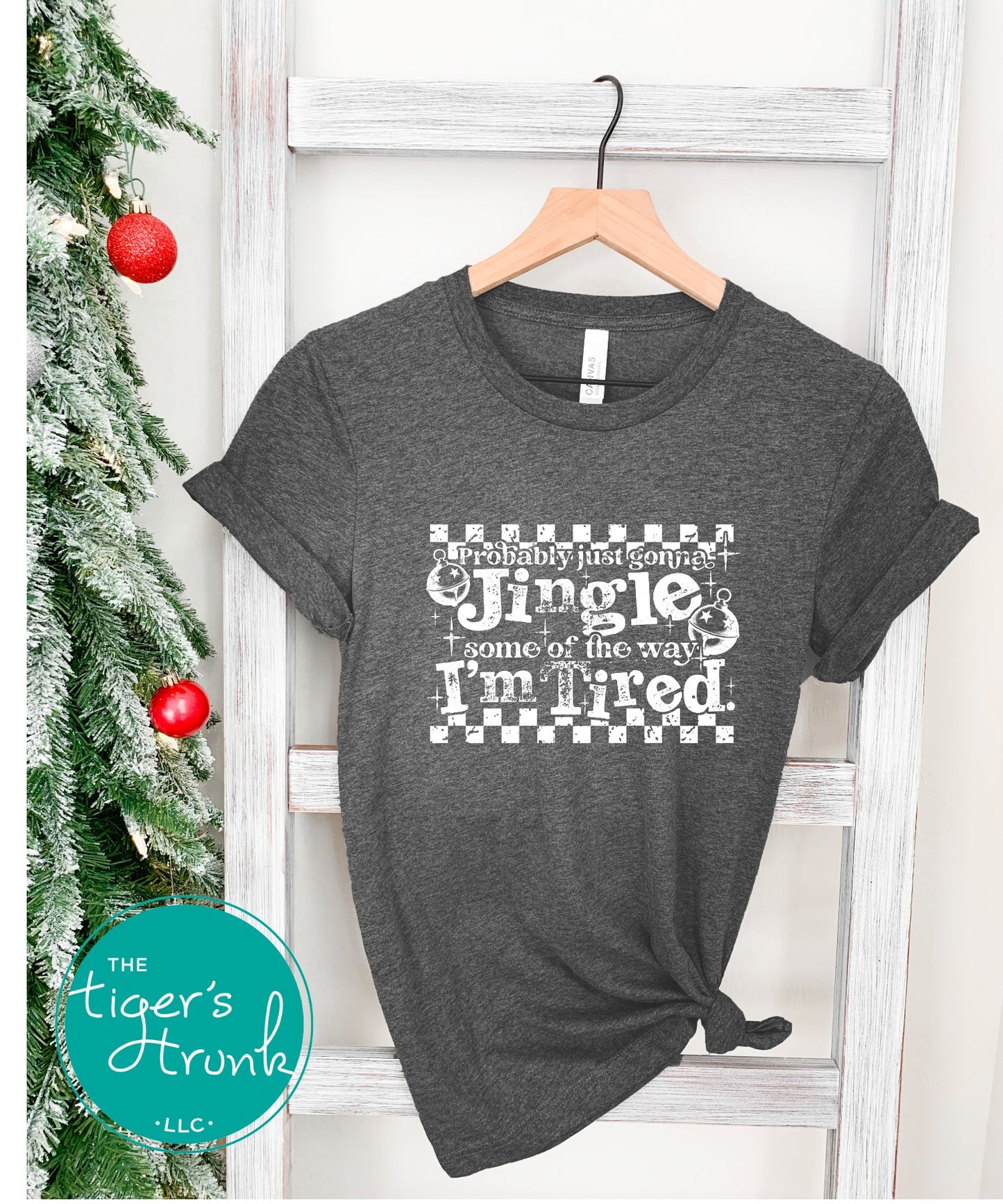 Gray t-shirt with Christmas-themed text design on a hanger in front of a decorated tree.