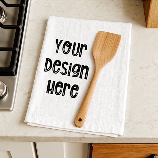 White towel with 'Your Design Here' text and wooden spatula on a kitchen counter.