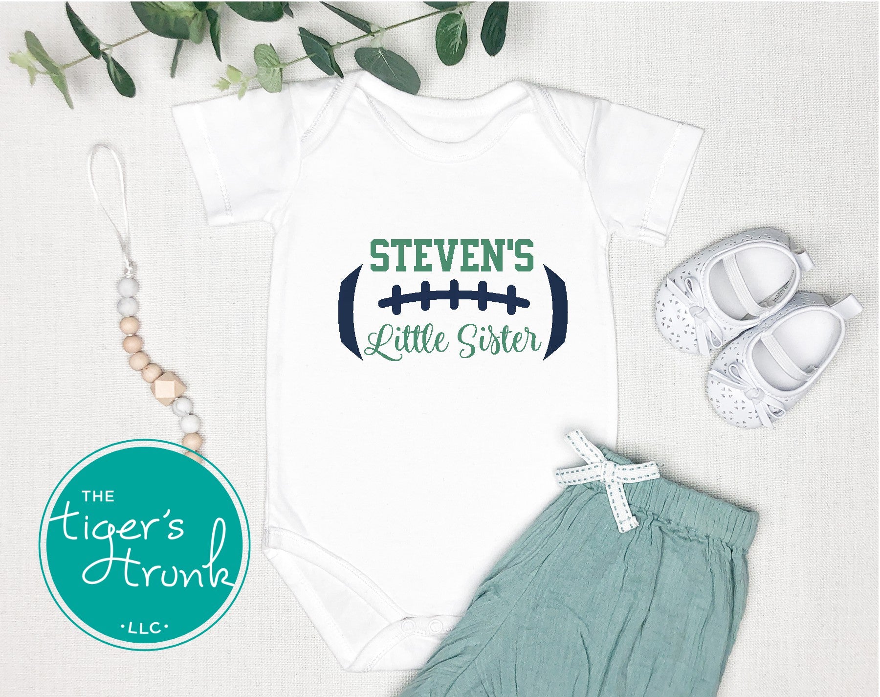 Baby onesie with 'Steven's Little Sister' text, baby shoes, and green pants on a light background.