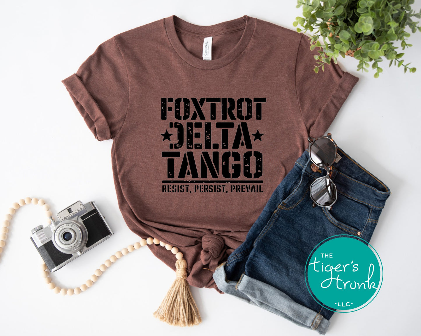 Brown t-shirt with 'Foxtrot Delta Tango' text, sunglasses, camera, and denim jeans on a white background.
