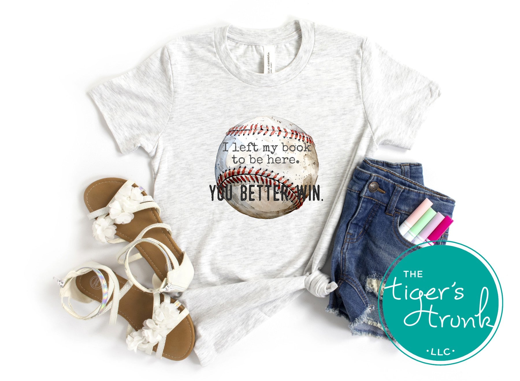 White t-shirt with baseball graphic and text, sandals, denim shorts, and stationery on a white background with 'The Tiger's Trunk' logo.