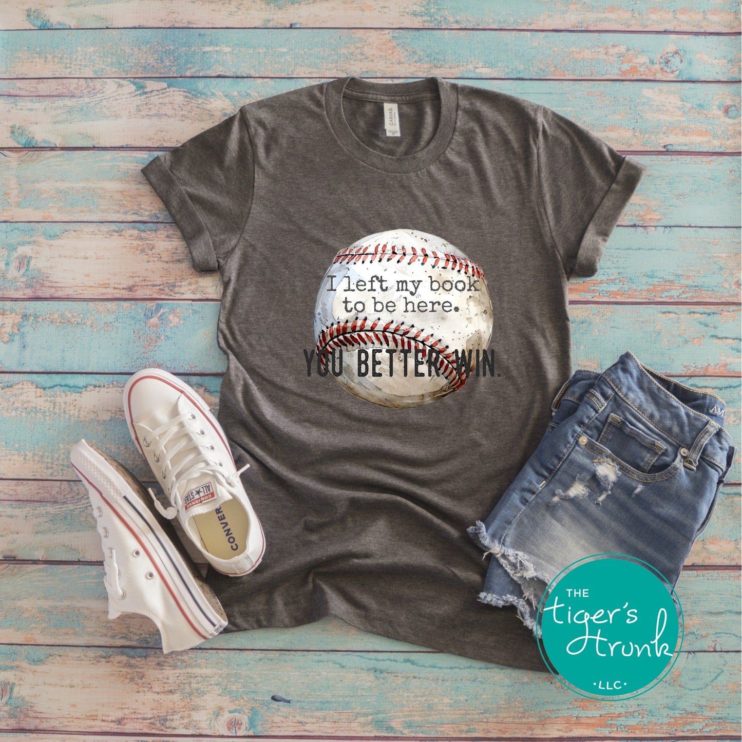 Gray t-shirt with baseball graphic and text, paired with jeans and sneakers on a wooden surface, branded 'The Tiger's Trunk'.