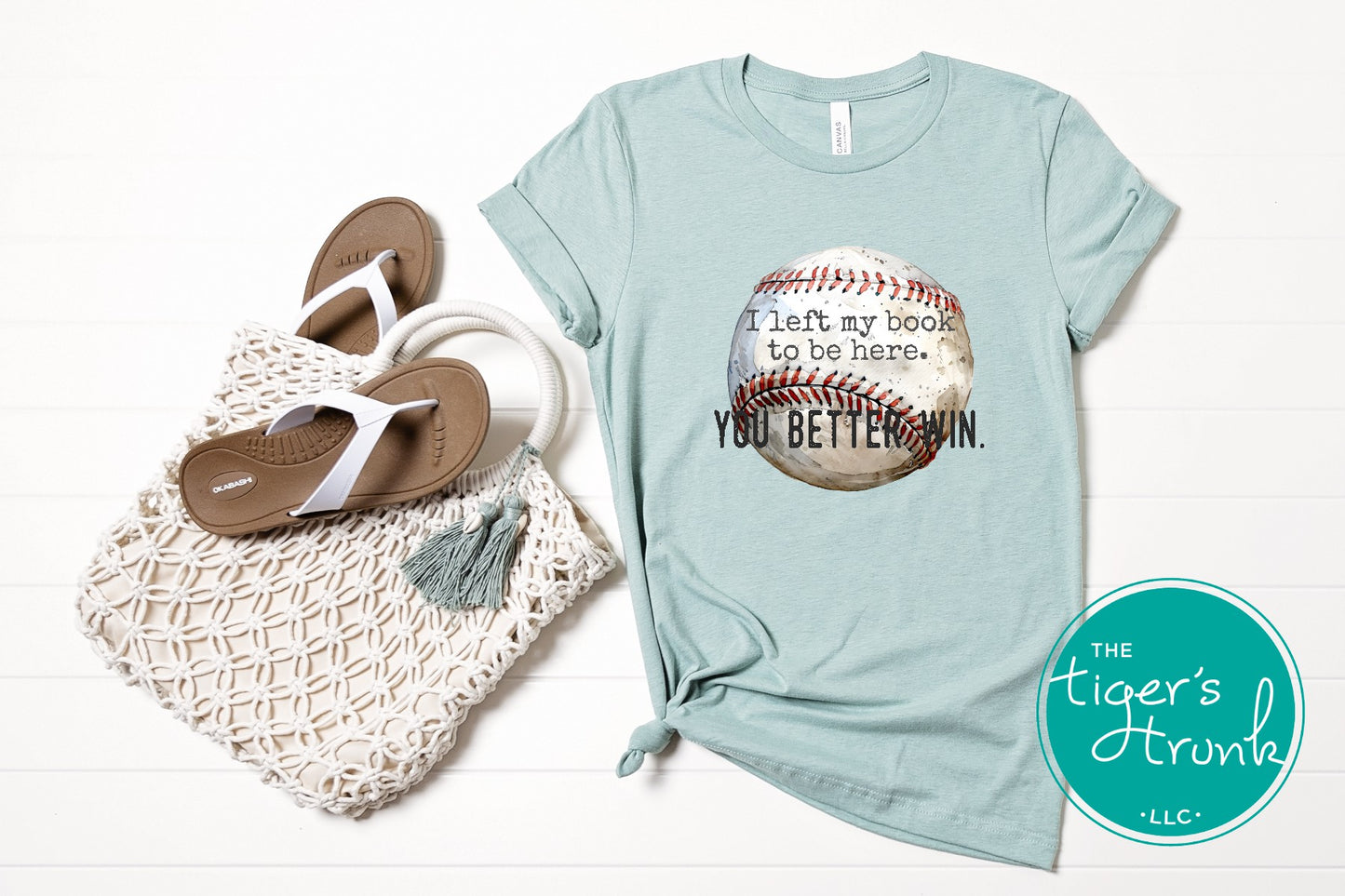 Light blue t-shirt with baseball graphic and text, next to flip-flops and a bag on a white background.