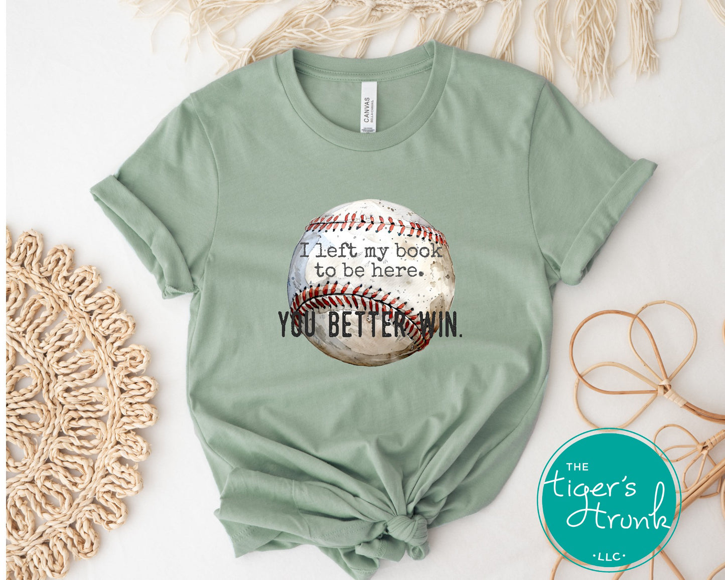 Sage green t-shirt with a baseball graphic and text on a white surface with decorative elements.