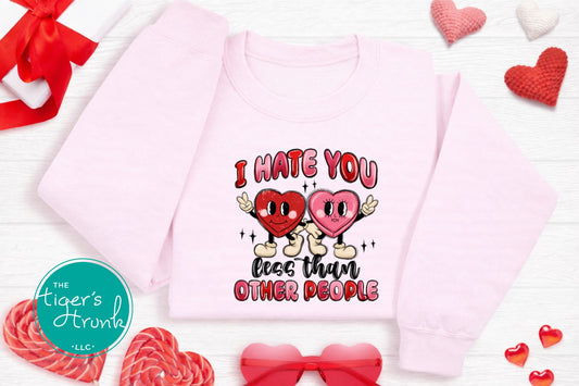 Pink sweatshirt with 'I Hate You Less Than Other People' text and hearts, surrounded by candy and bows on a white background.