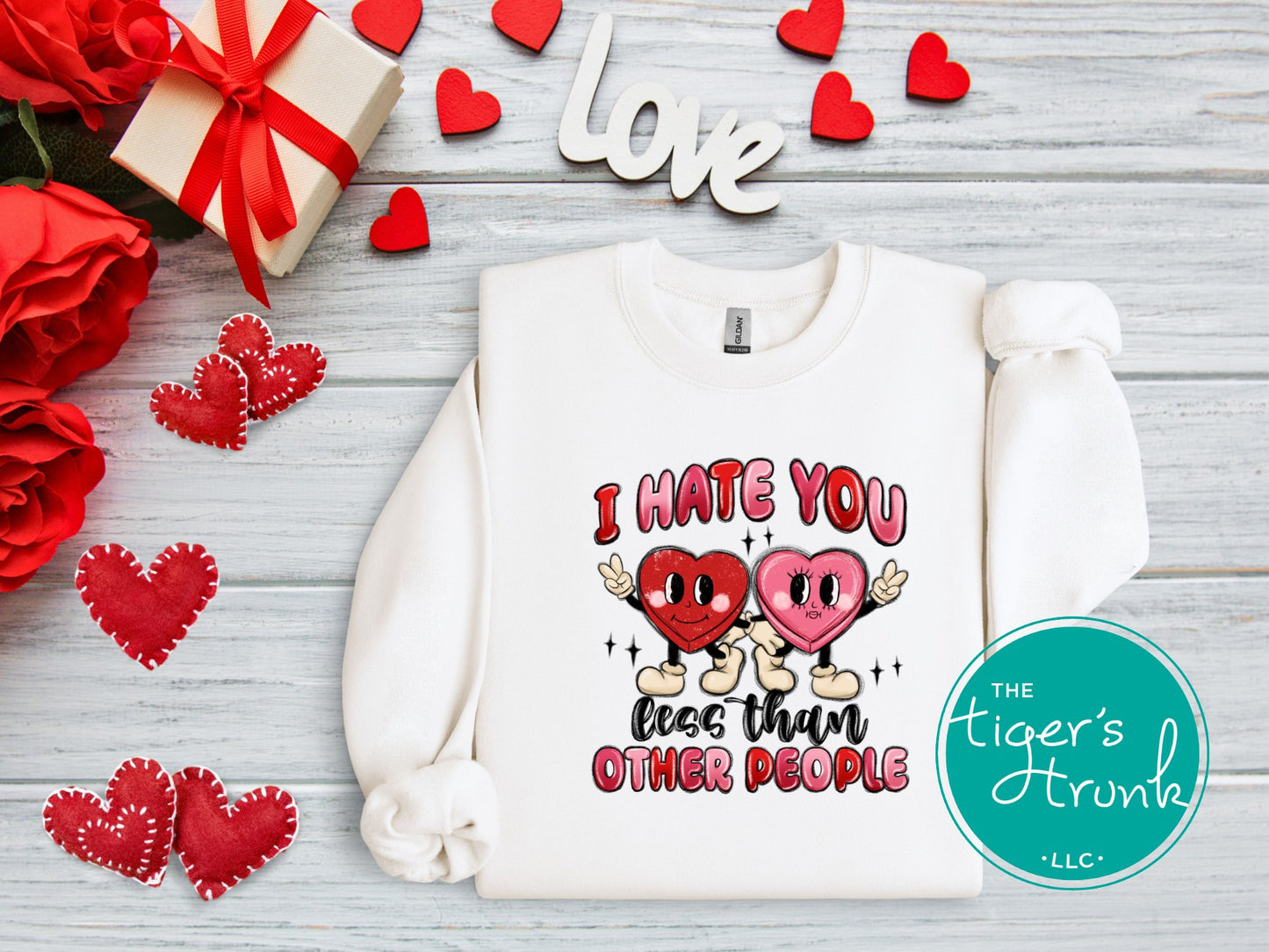 White sweatshirt with graphic design and text, surrounded by red hearts, roses, and a gift box on a wooden surface.