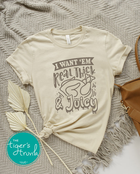 A short-sleeve beige shirt with the text 'I WANT 'EM REAL THICK & JUICY' printed on the front, displayed on a grey and white faux fur blanket with a decorative feather and a leather-look belt.