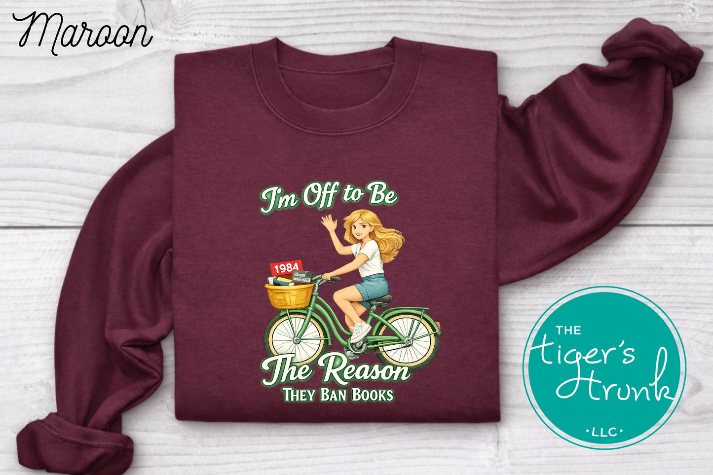 Maroon progressive sweatshirt featuring a reader riding a bicycle with banned books and the phrase I’m off to be the reason they ban books