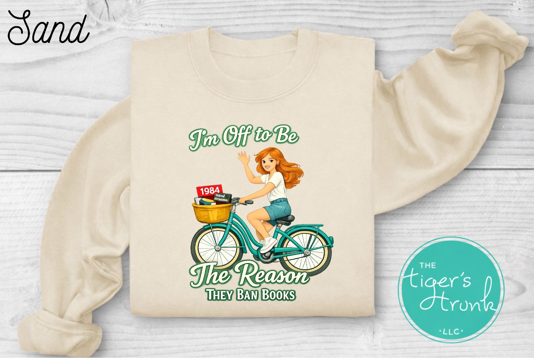 Sand colored progressive sweatshirt featuring a reader riding a bicycle with banned books and the phrase I’m off to be the reason they ban books
