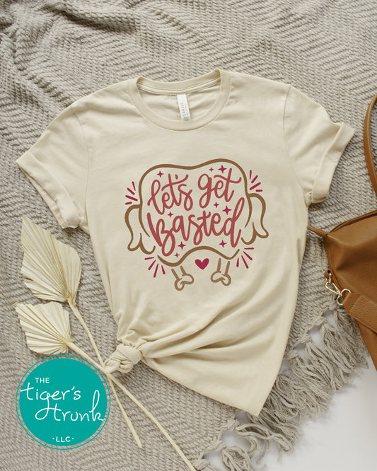 A short-sleeve beige thanksgiving themed shirt with the text 'Let's Get Basted' printed on the front, displayed on a grey, fringed blanket with a white turkey decoration and a feather beside it.