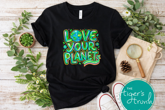 Black t-shirt with 'Love Your Planet' design on a wooden surface with greenery and a magnifying glass.