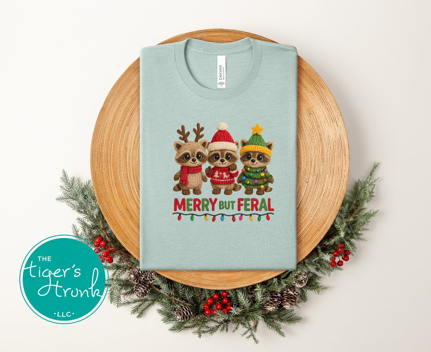 Light blue t-shirt with Christmas-themed design of three animals wearing hats and scarves, surrounded by a decorative wreath.