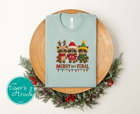 Light blue t-shirt with Christmas-themed design of three animals wearing hats and scarves, surrounded by a decorative wreath.