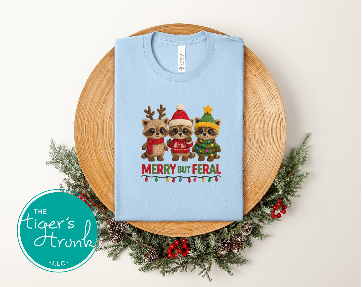Light blue t-shirt with Christmas-themed raccoon design on a wooden plate with greenery