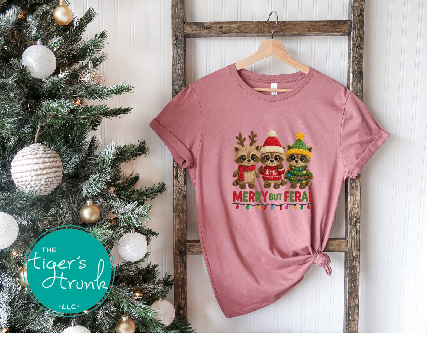 Pink t-shirt with Christmas-themed design of two animals and text, hanging on a wooden rack next to a decorated Christmas tree.