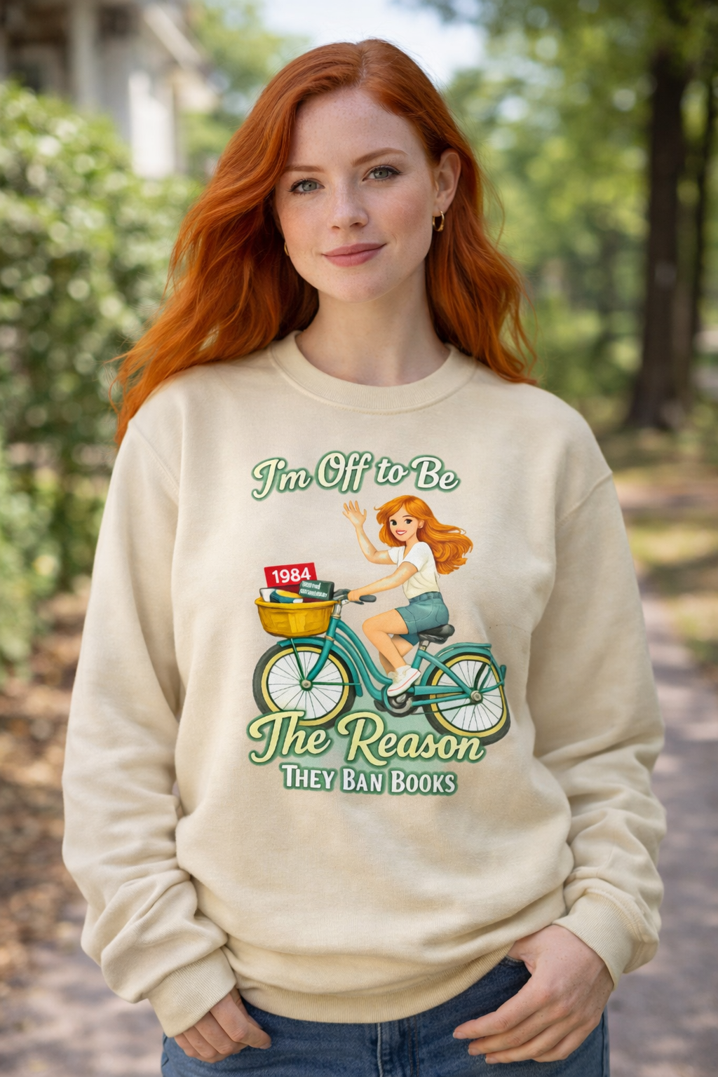 Red-haired woman wearing a sand colored progressive sweatshirt that says I’m off to be the reason they ban books with a banned books bicycle graphic