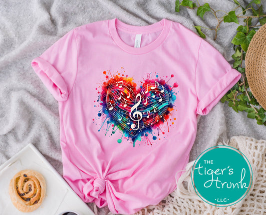 Pink t-shirt with colorful heart and music notes design on a textured surface with 'The Tiger's Trunk' logo.