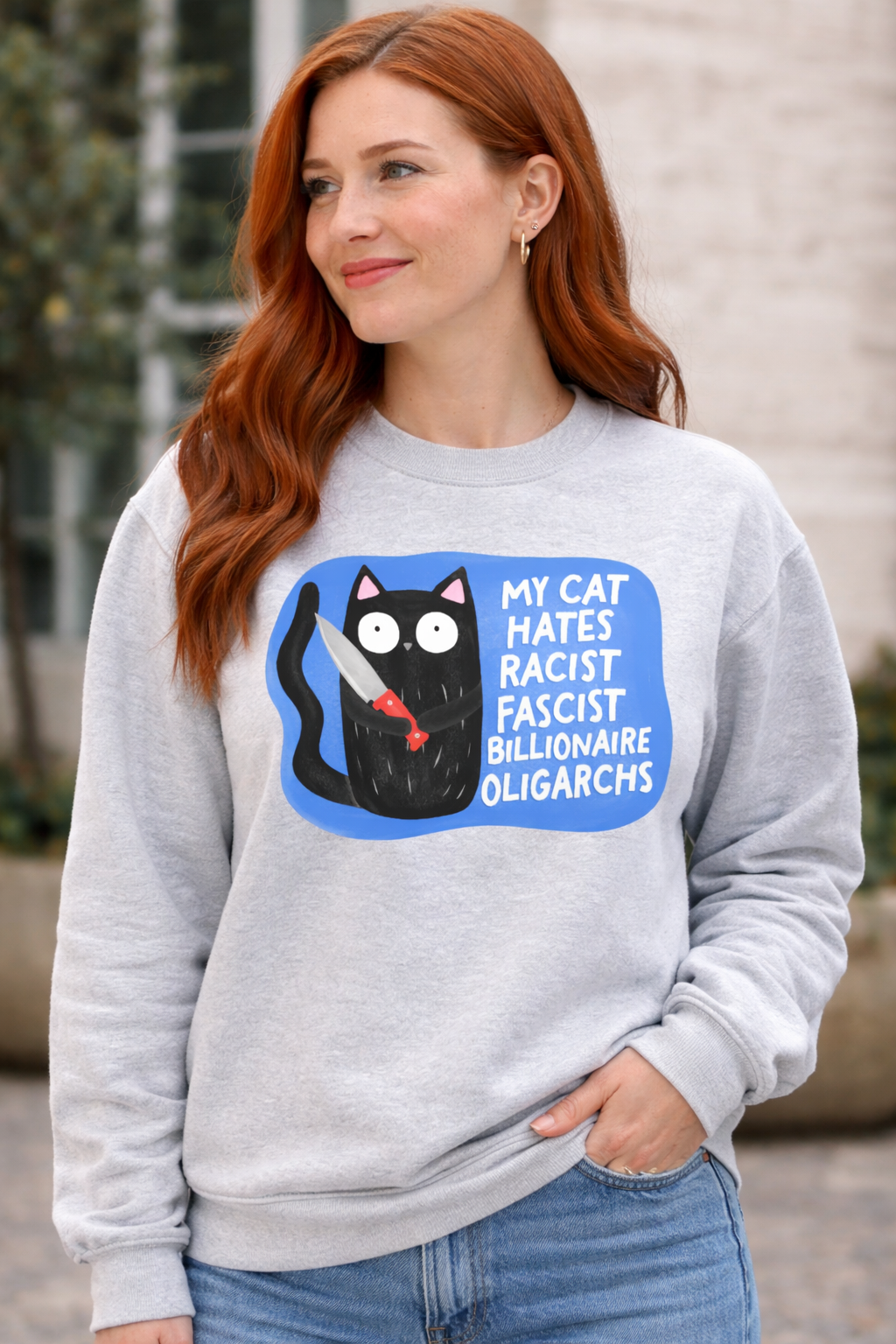 Red-haired woman wearing an ash gray sweatshirt featuring a black cat graphic with the phrase My Cat Hates Racist Fascist Billionaire Oligarchs, political protest crewneck