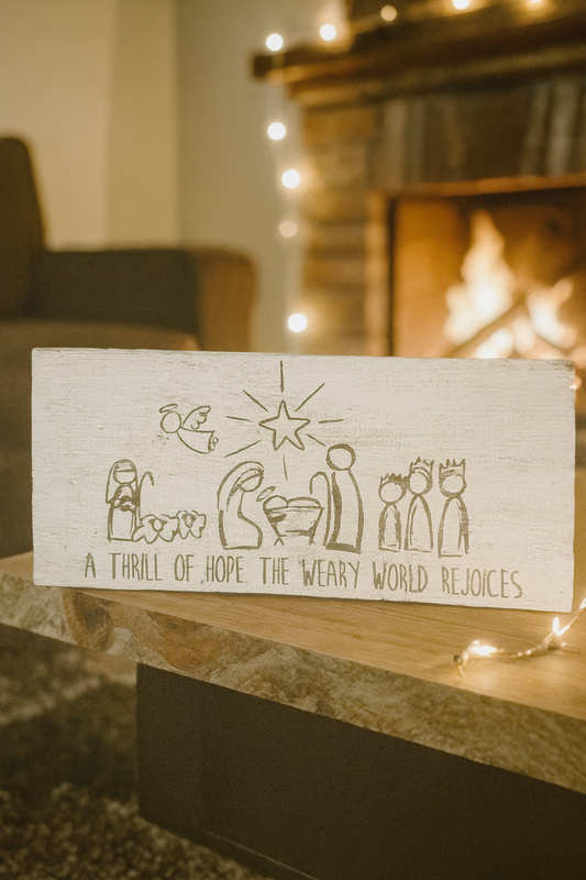 Hand-painted wooden nativity sign displayed on a mantel with soft Christmas lights.