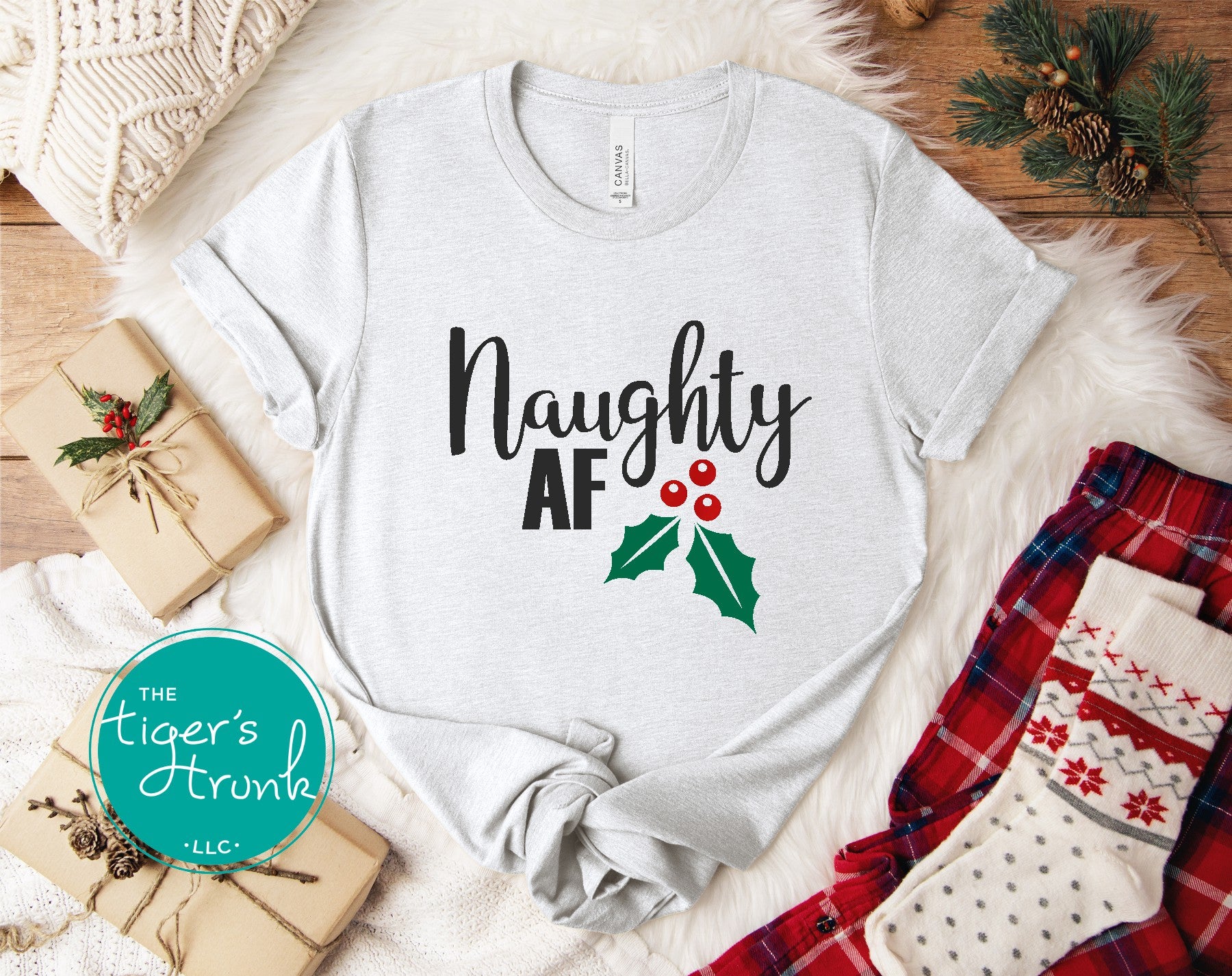 Gray t-shirt with 'Naughty AF' text and holly design on a festive background with presents and plaid fabric.