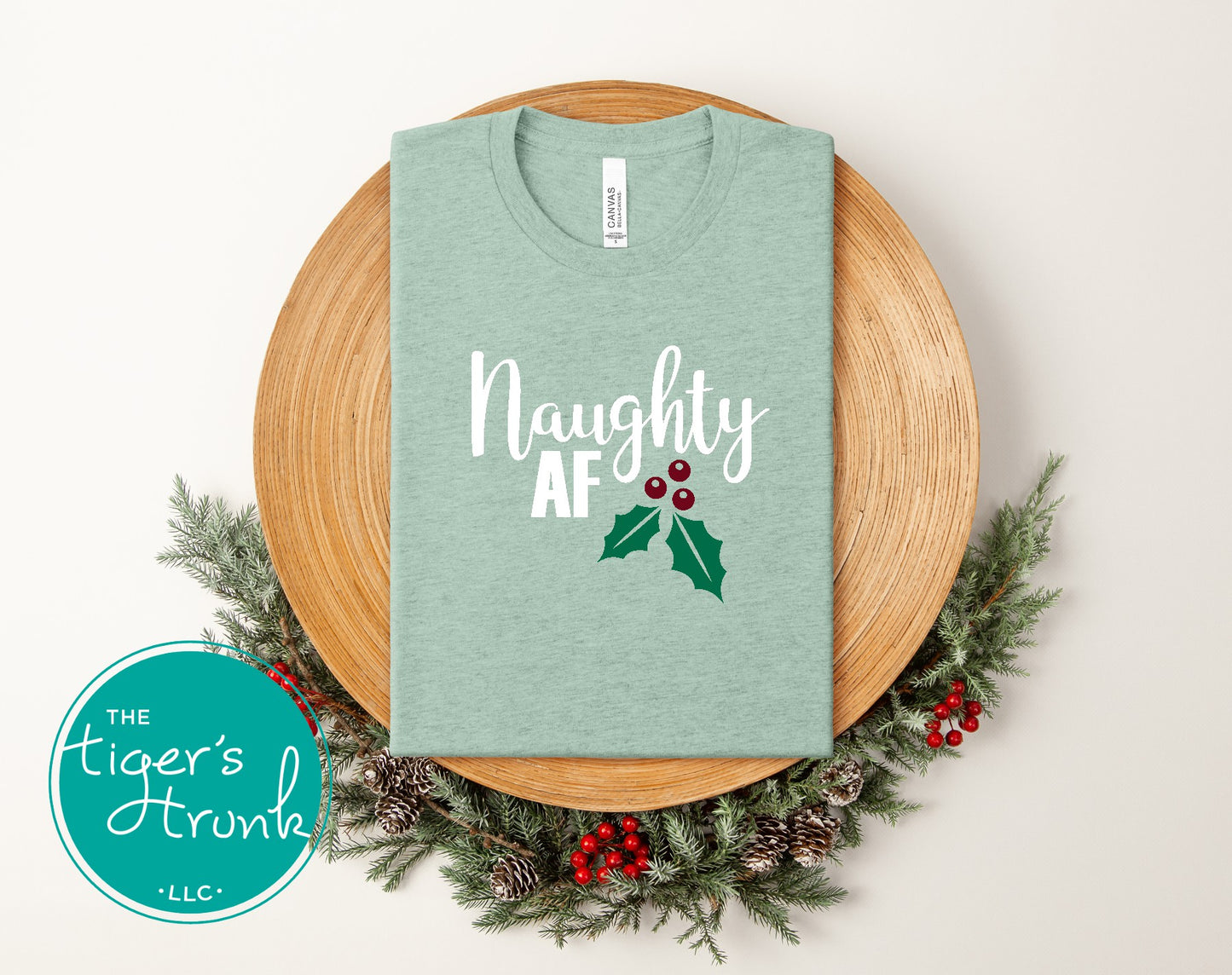 Green t-shirt with 'Naughty AF' text on a wooden plate with Christmas decorations.