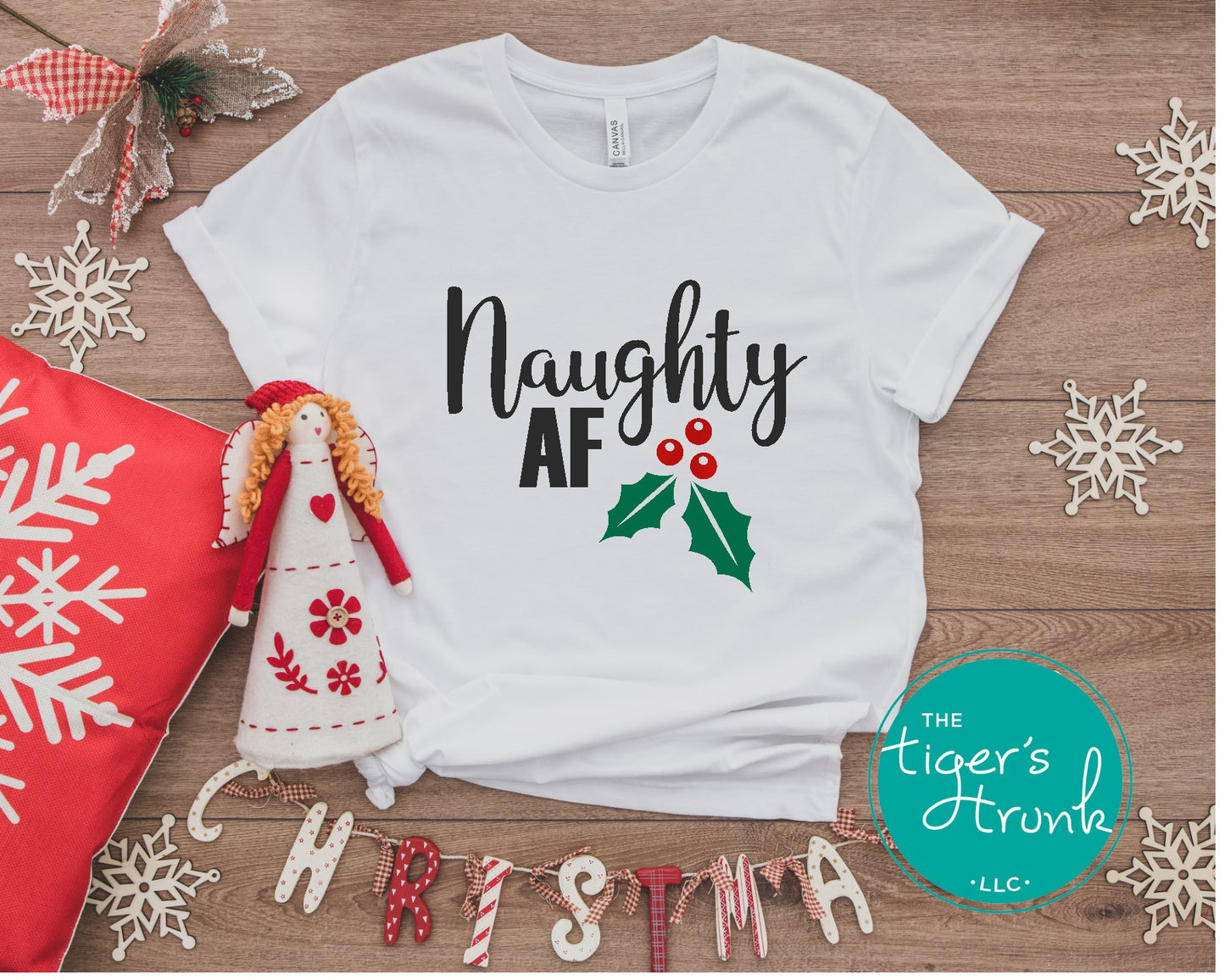 White t-shirt with 'Naughty AF' text and holly design on a wooden surface with Christmas decorations.