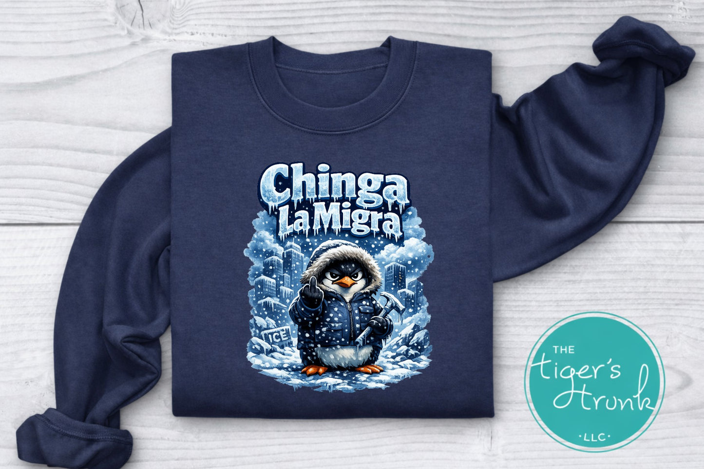 Navy blue sweatshirt with a penguin graphic and 'Chinga La Migra' text on a light wooden surface, with 'The Tiger's Trunk' logo in the corner.