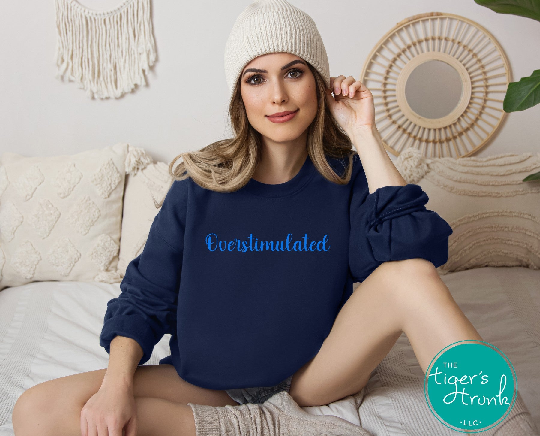 Model wearing navy blue overstimulated sweatshirt with blue script text sitting on bed in cozy neutral setting
