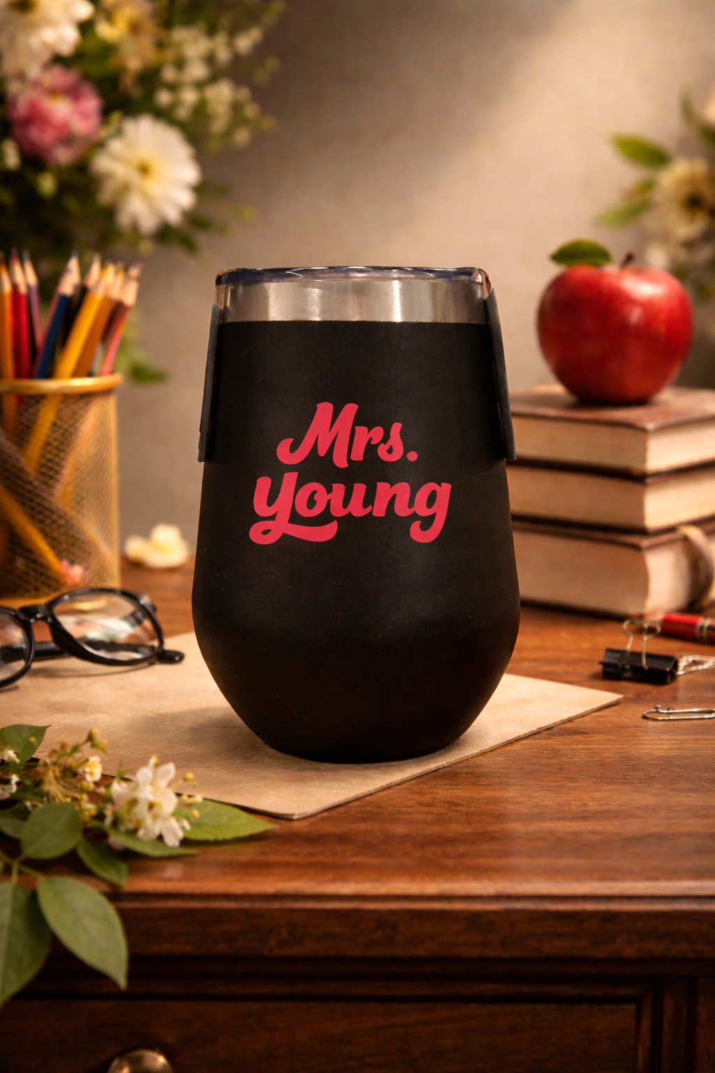 Black tumbler with 'Mrs. Young' in red text on a wooden desk with flowers and books in the background.