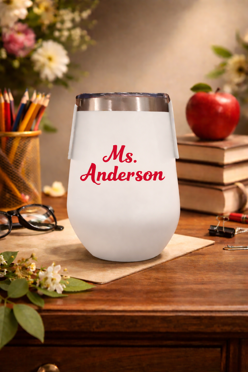 White tumbler with 'Ms. Anderson' printed on it, placed on a wooden surface with books and flowers in the background.