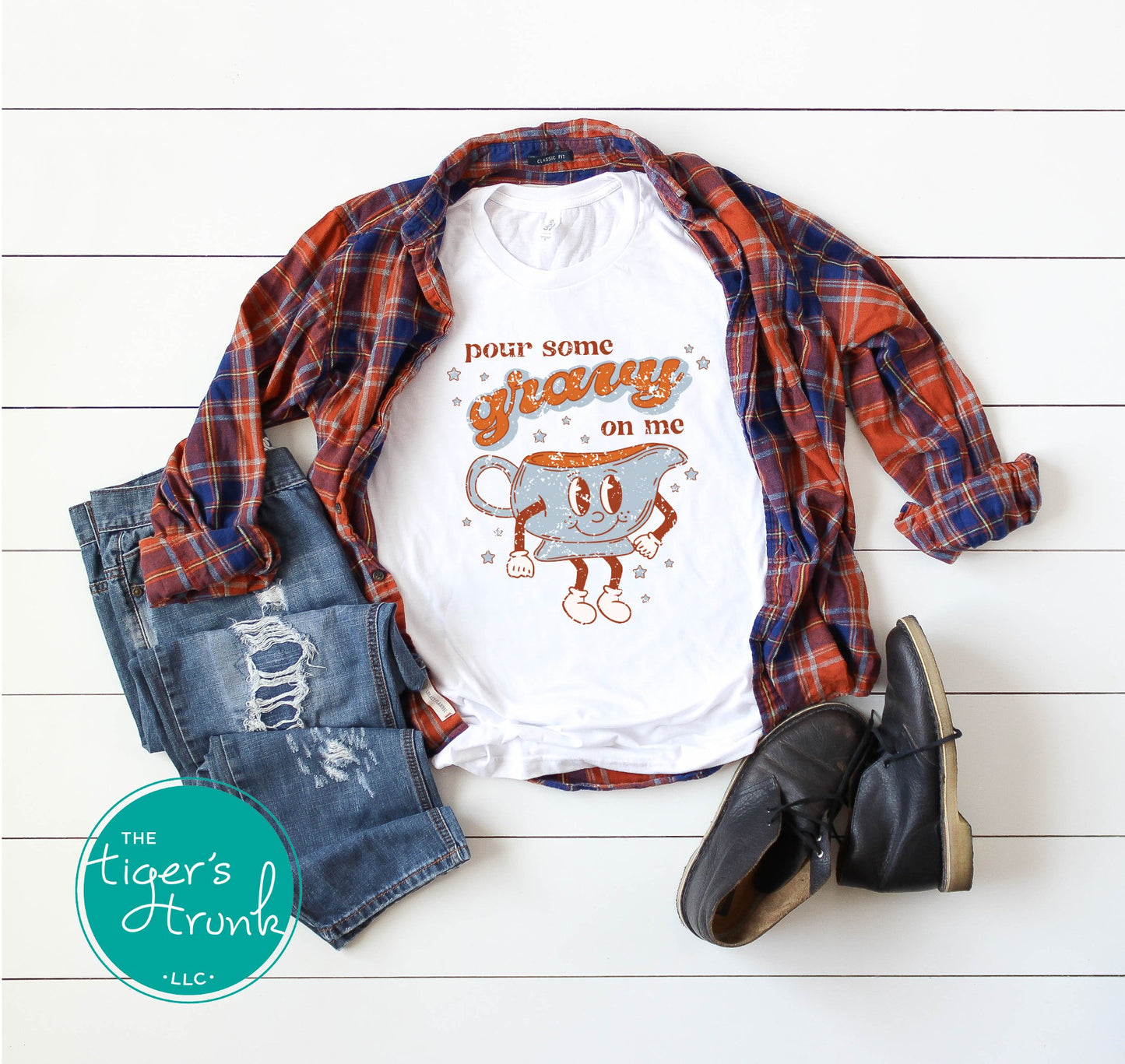White t-shirt with graphic design, plaid shirt, jeans, and boots on a white wooden surface with 'The Tiger's Trunk' logo.