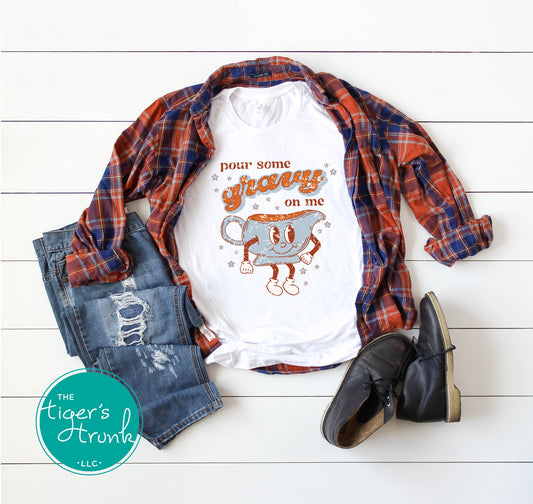 White t-shirt with graphic design, plaid shirt, jeans, and boots on a white wooden surface with 'The Tiger's Trunk' logo.