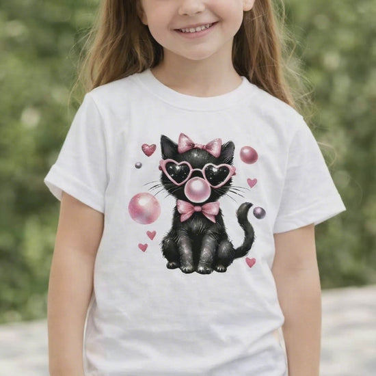 Child wearing a white short sleeve t shirt featuring a black cat Valentine design with pink heart sunglasses, bow, and floating heart accents, shown on a youth model outdoors