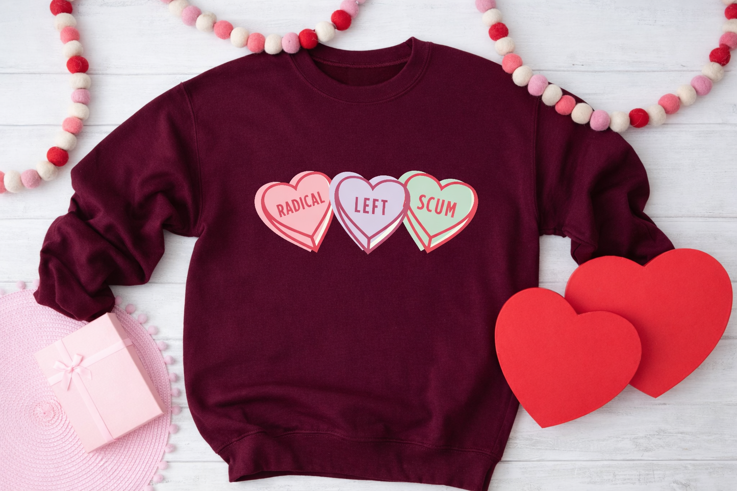Maroon sweatshirt with heart-shaped text surrounded by decorative hearts and a garland on a light wooden surface.